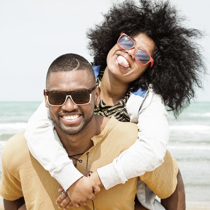 Young afro american couple playing piggyback ride on beach - Che